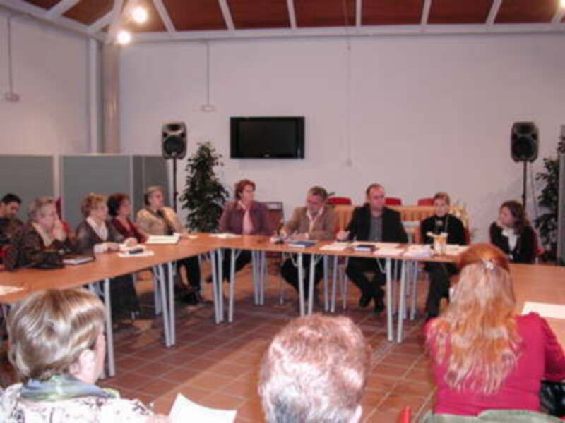 Una reunión formal en una sala con mesas redondas, personas sentadas y un televisor en el fondo. La imagen muestra una reunión de trabajo o conferencia con participantes sentados y un público observando.
