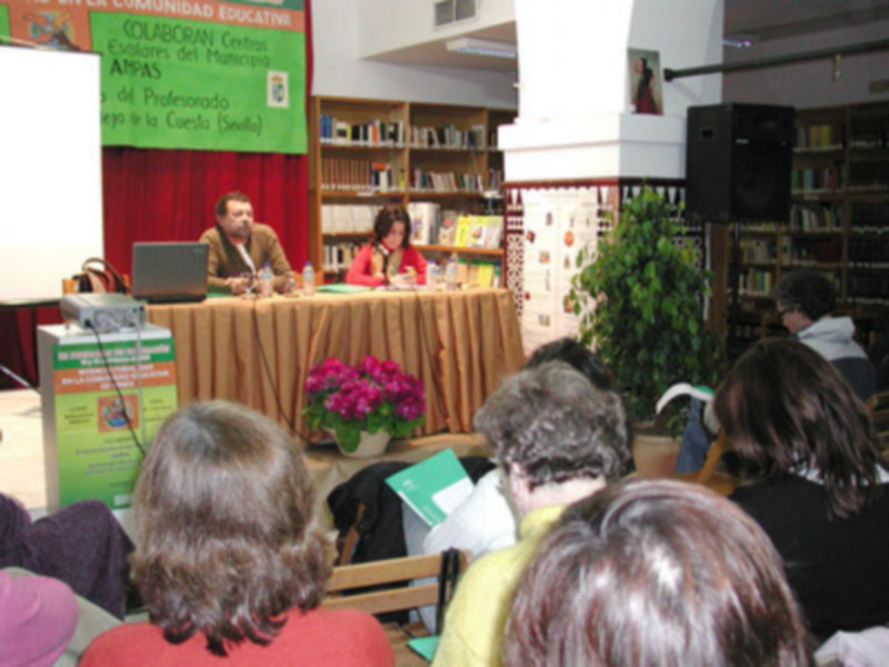 Una conferencia en un salón de conferencias con una mesa central, donde dos personas están sentadas. En la pared hay libros y un cartel informativo sobre la Comunidad Educativa. La audiencia está sentada en filas, escuchando atentamente.