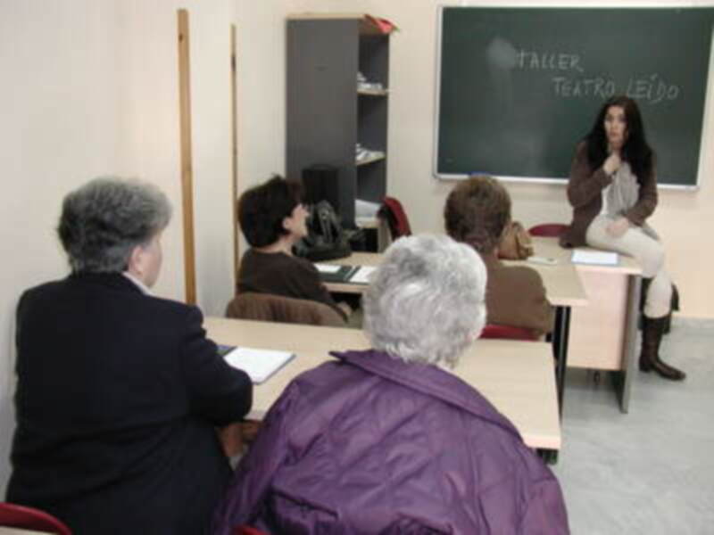 Taller de teatro dirigido por una mujer sentada en la mesa. La clase está formada por personas mayores sentadas en sillas frente a una pizarra con la palabra "Taller Teatro Leído".