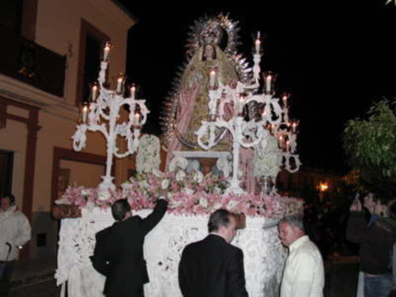 Una procesión religiosa con una imagen de la Virgen en un carro adornado con flores y velas, acompañada por fieles.