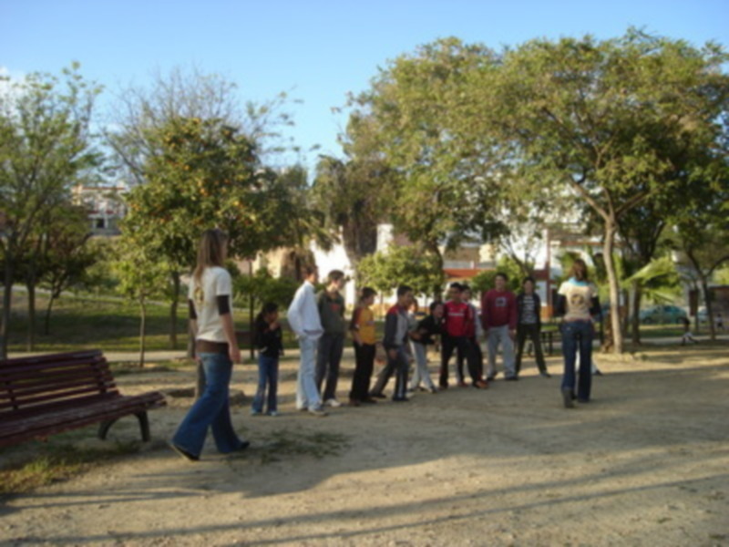 Un grupo de personas en un parque, con árboles y una casa al fondo. La gente está de pie en fila, algunas personas están sentadas en un banco. El cielo es claro y el sol brilla.