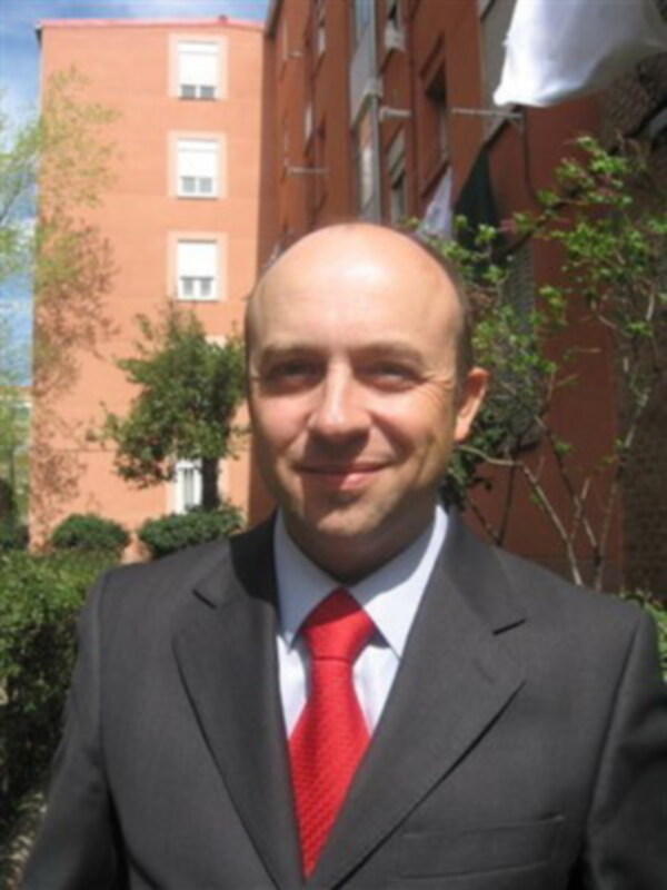 Un hombre con una camisa blanca, corbata roja y traje negro. Está en un entorno urbano con edificios de fondo, un árbol y cielo azul.
