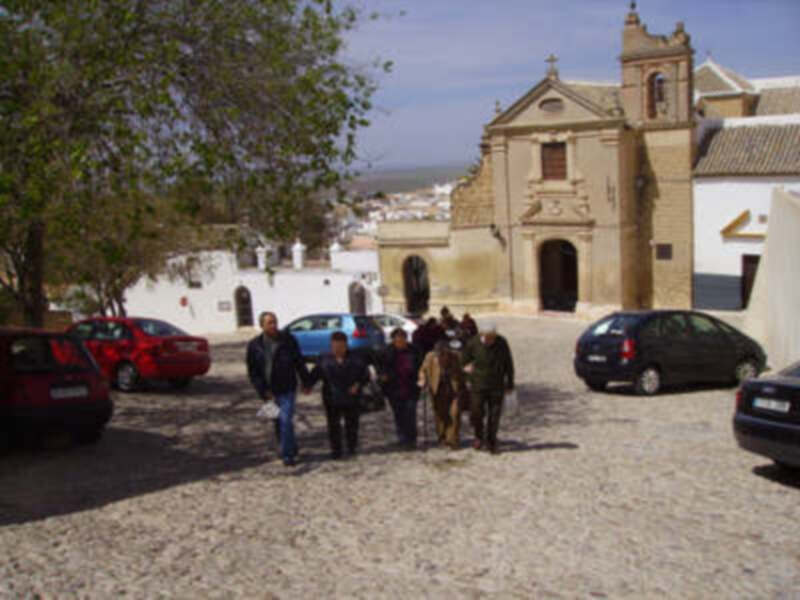 Un grupo de personas caminan por una calle adoquinada, con edificios históricos a ambos lados. En el fondo se observa un coche azul y varios vehículos estacionados, junto a una iglesia con detalles arquitectónicos.