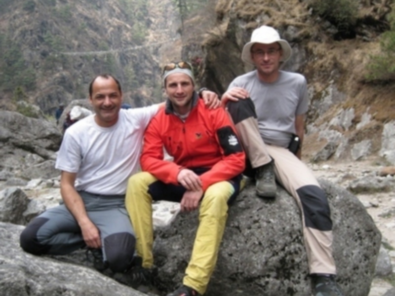 Tres hombres sentados en una roca con paisaje montañoso y cascada al fondo.
