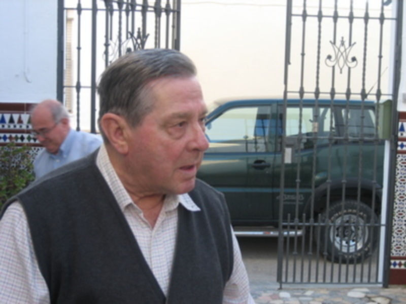 Hombre mayor con chaleco negro frente a una verja de hierro forjado y un vehículo verde en el fondo.