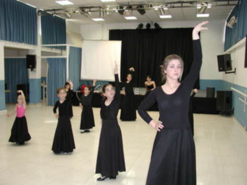 Las bailarinas en el escenario están ejecutando una coreografía sincronizada, con vestimentas negras y cabellos sueltos. La escena transmite un ambiente de práctica artística en una sala de baile con cortinas azules y luces iluminando el espacio.