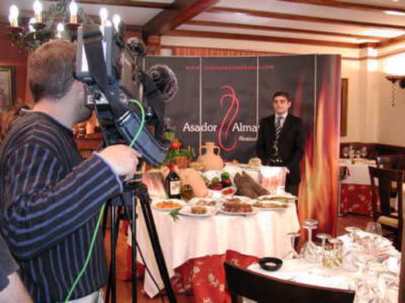 En un restaurante, un cameraman filma a una persona vestida de negro frente a un cartel con el nombre "Asador Alma". La mesa está servida con platos y copas.