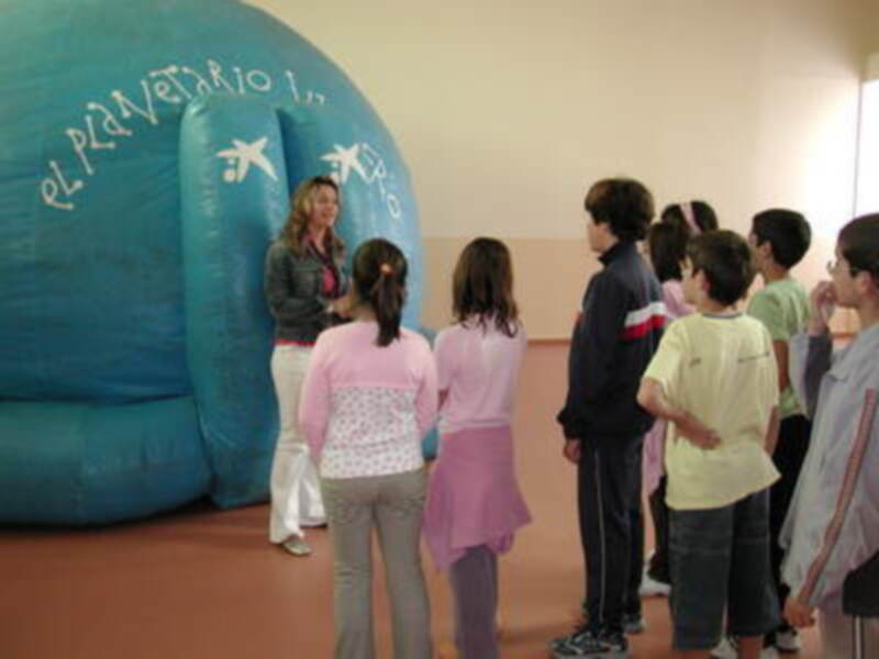 Una niña con un vestido rosa y una falda azul está en el centro de la imagen, rodeada por otros niños. Al fondo, una mujer habla con un grupo de niños en un espacio inflable azul con el texto "el Planetario".
