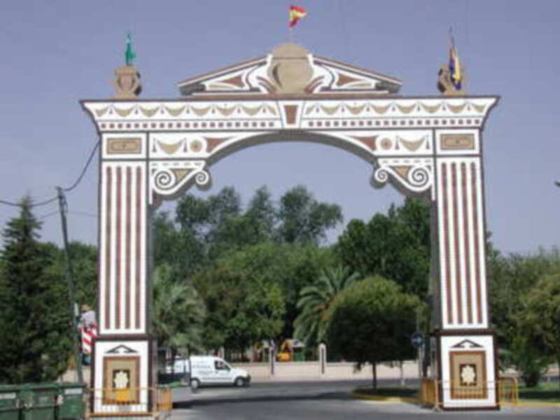 Puerta monumental con detalles clásicos y banderas ondeando, frente a un camino con árboles verdes.