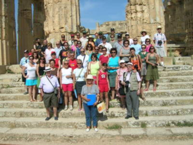 Un grupo de turistas en un parque histórico, posando para una foto frente a una estructura antigua. El cielo está despejado y el ambiente es soleado, indicando un día perfecto para visitar el lugar.