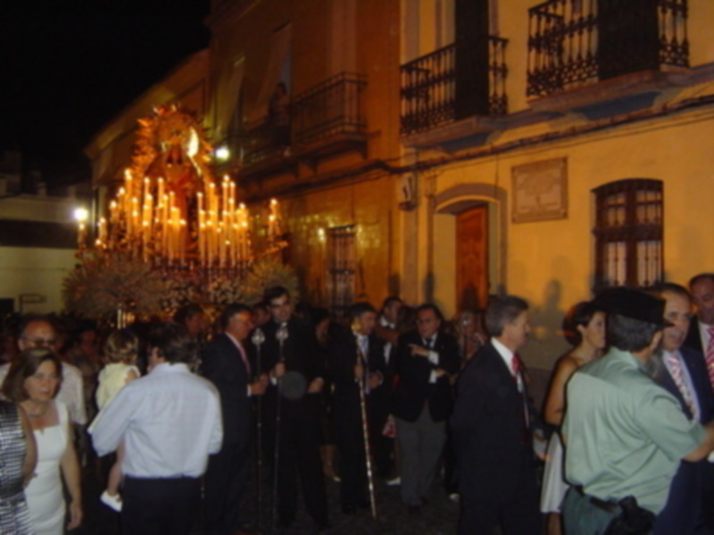 Una procesión nocturna con personas vestidas formalmente, rodeadas de luces y decoraciones festivas en un edificio histórico.