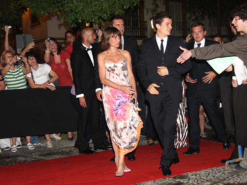 Persona en vestido floral y hombre en traje negro caminando por alfombra roja en evento formal.