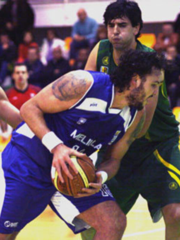 Un jugador de baloncesto en azul y blanco intenta controlar el balón mientras otros jugadores lo persiguen. La escena se desarrolla en un estadio lleno de espectadores.