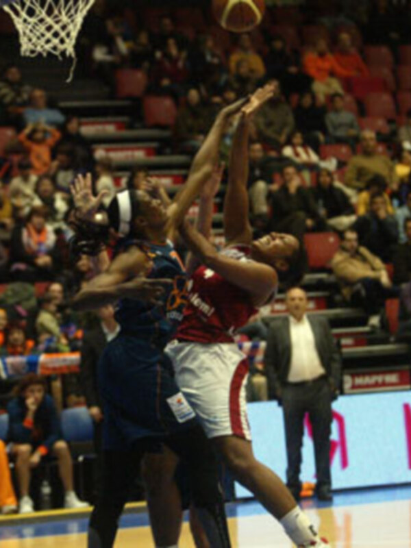 Un jugador de baloncesto rojo y negro intenta anotar mientras otro jugador azul lo bloquea en un partido de baloncesto. La arena está llena de espectadores y el fondo muestra una pantalla publicitaria con el número 1.