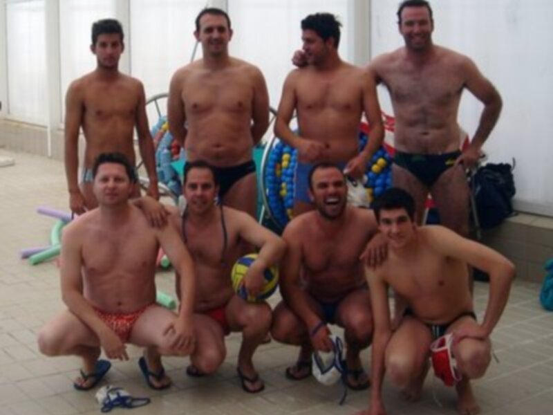 Un grupo de hombres en trajes de baño posan juntos en una piscina. Algunos llevan pelotas y otros están con sus manos en el aire, posiblemente celebrando. La escena parece ser una reunión o un evento deportivo en el interior de la piscina.