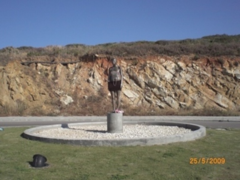 Una estatua de bronce en un pedestal en el centro de una rotonda de piedra, rodeada por vegetación y con un fondo de acantilado rocoso. La fecha en la imagen es 25/5/2009.