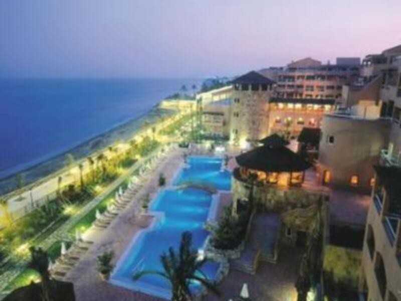 Vista nocturna de un hotel con piscina y palmeras, ubicado junto al mar.