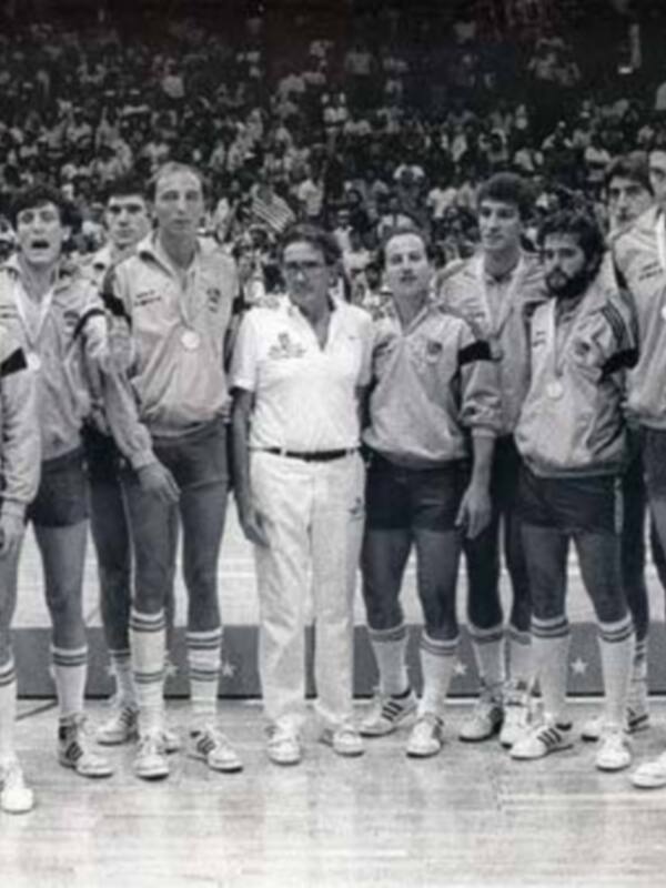 Equipo de baloncesto con entrenador y medallas, posando en un estadio lleno de espectadores.
