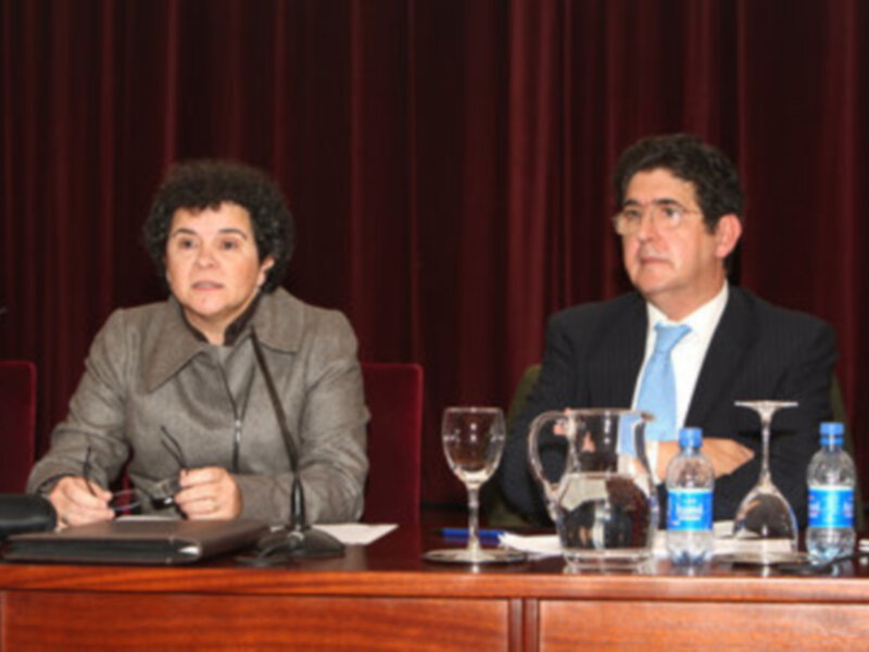 Conferencia en sala formal con dos personas sentadas en un panel, rodeadas de mesas con agua y vajilla.