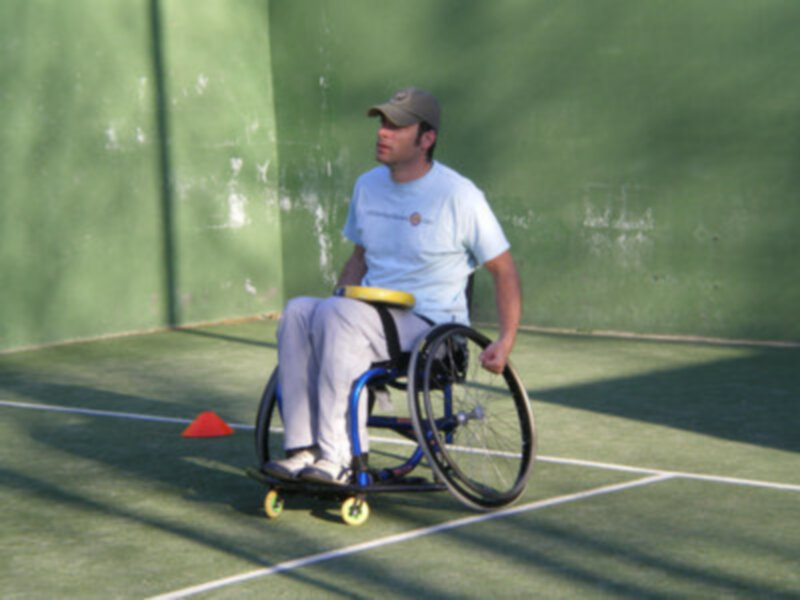 Un hombre en silla de ruedas con una gorra gris y camiseta azul está sentado en un tenis court. La imagen muestra su silla de ruedas con ruedas amarillas y una cancha verde.