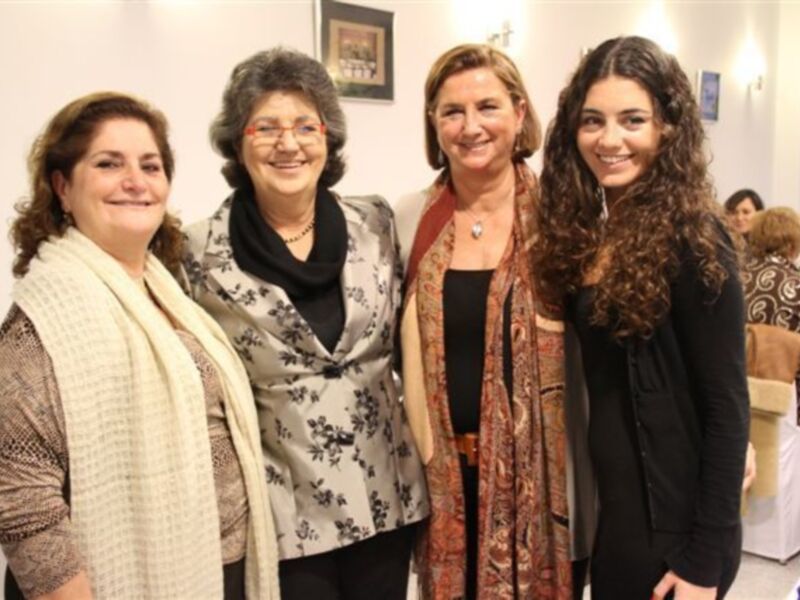Cuatro mujeres sonríen y posan juntas en un evento social, rodeadas de decoración elegante y otras personas en el fondo.