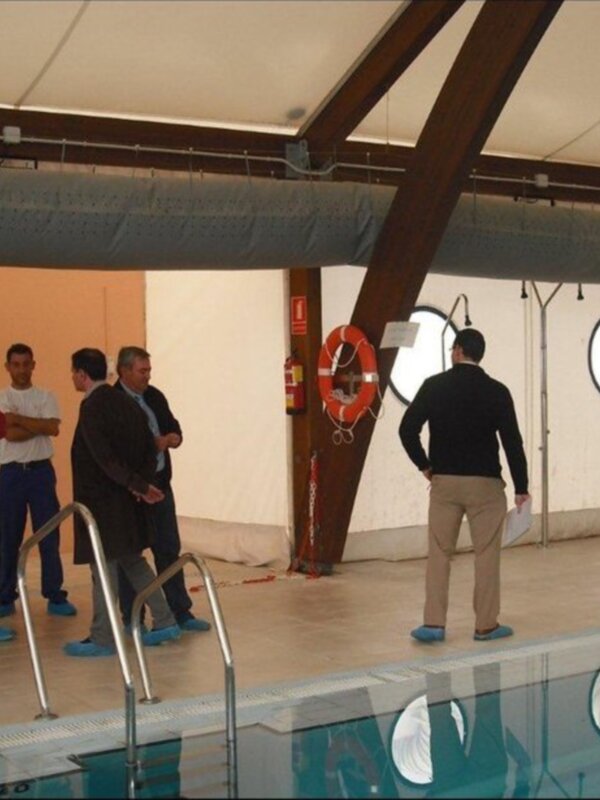 En una piscina, un grupo de personas observa a un hombre con un traje de baño y una mochila. A la derecha, hay un resbalón y un salvavidas colocado en el muro.