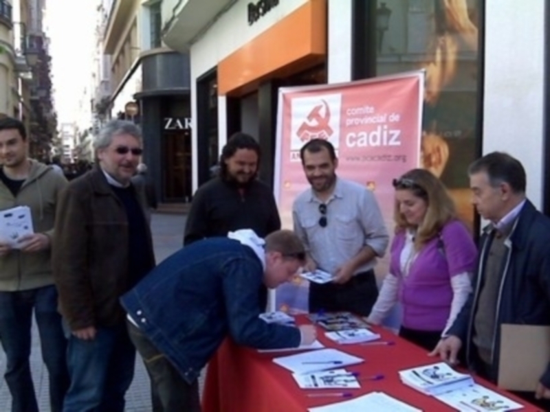 Una persona firmando un documento en una mesa roja, con personas observando y sosteniendo folletos en la calle.