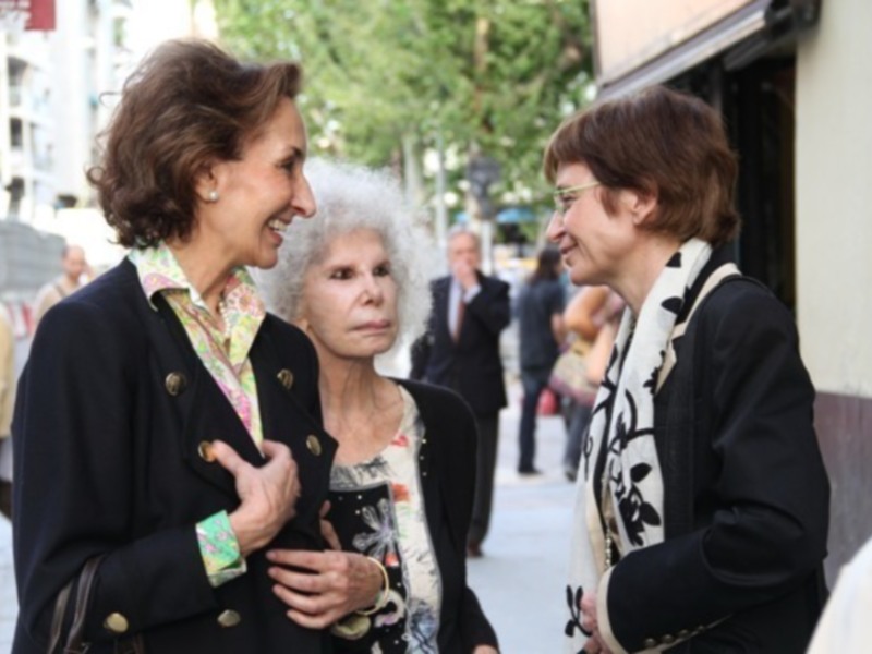 Tres mujeres intercambian sonrisas en una calle urbana, rodeadas de gente y edificios.