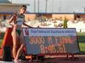 Una atleta femenina en ropa deportiva verde y roja, con el nombre Augusto, corre por una pista de atletismo. En primer plano se ve un tablero electrónico que muestra "3000 M FINAL" y el tiempo 8:46.51, junto con la marca "Real Federación Española de Atletismo" y el nombre "TEXTEL".