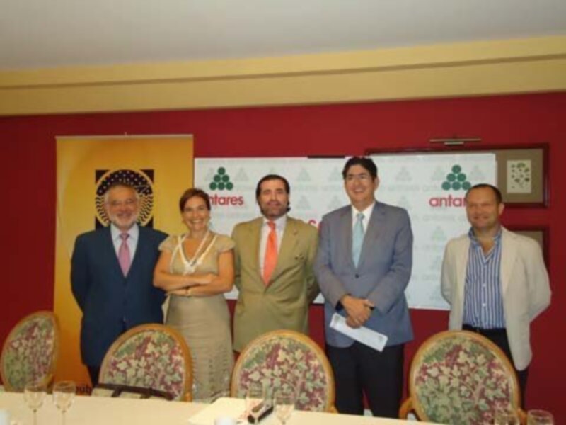 Una reunión formal con cuatro personas posando frente a una pantalla con logos de Antares. La escena es en un ambiente formal, con sillas tapizadas y un ambiente elegante.