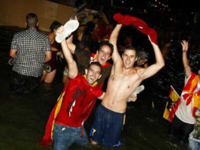 Fotografía de una celebración festiva en un ambiente nocturno. Personas vestidas con camisetas y banderas ondeando, algunos con pantalones cortos. La celebración parece estar en un espacio al aire libre con iluminación de fondo.