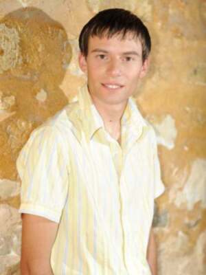 Un hombre joven con cabello corto y una camisa de manga corta en color amarillo. Fondo de pared de ladrillos antiguos.