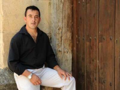 Un hombre con camisa negra y pantalones blancos se sienta en la entrada de una casa con una puerta de madera.