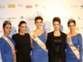 Las cinco mujeres posan frente a un fondo con logos de diferentes empresas. La mujer en el centro lleva una corona y un cinturón con la palabra "Miss" seguida de números. A su izquierda, una mujer con un cinturón azul y a la derecha, otra con un cinturón rojo.