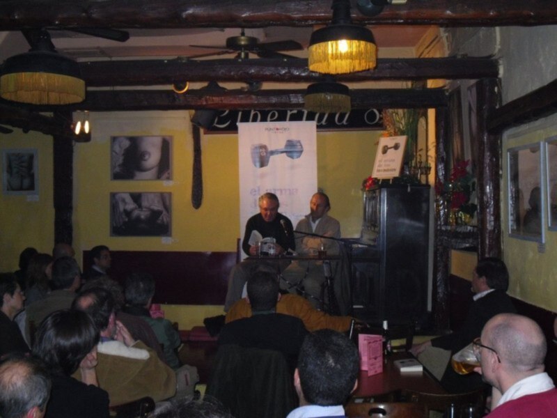 Lectura en una cafetería acogedora con público atento y carteles de eventos en el fondo.