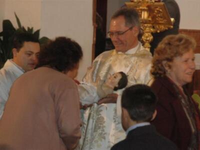 Una imagen de una ceremonia religiosa donde un sacerdote sostiene a un niño en sus brazos, rodeado por personas que observan la escena. La atmósfera es solemnemente reverente, con elementos decorativos y una pila bautismal en el fondo.