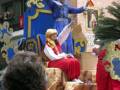 Imagen de un desfile navideño con una figura vestida como un rey en el centro, rodeada de personas y decoraciones navideñas.