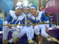 Tres jóvenes vestidos de azul y blanco participan en un desfile, con sombreros y accesorios dorados.