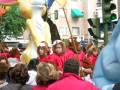 Desfile infantil con niños vestidos de personajes de dibujos animados, entre ellos un oso y un dinosaurio. El público observa la celebración en una calle con edificios a lo lejos y un semáforo visible.