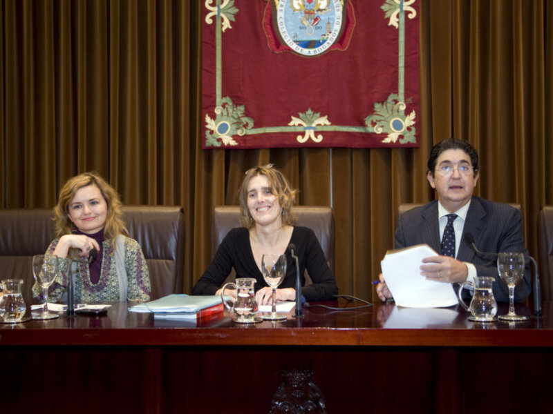 Tres personas sentadas en una mesa con documentos y copas de vino, ante un escudo de armas en la pared.