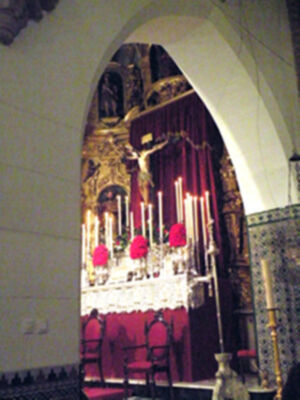 Interior de una iglesia con un altar adornado con velas y una imagen de Cristo crucificado en el centro. El arco del altar está cubierto con velas encendidas y sillas rojas alrededor.