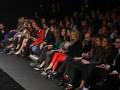 Una audiencia de personas sentadas en un evento, algunos con zapatos de tacón alto y otros con zapatos planos, en un escenario oscuro con luces de fondo.