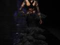Una modelo camina por la pasarela de una moda, con un vestido negro de encaje y volantes, llevando flores en la espalda. La iluminación es dramática y el fondo es oscuro, enfocando la atención en el vestido y las manos de la modelo.