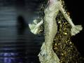 Una modelo vestida con un vestido de gala dorado y brillante, con detalles florales, camina por un pasarela iluminada por fuegos artificiales en el fondo.
