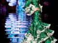 Una mujer vestida de verde con detalles blancos camina por un pasarela iluminada, su vestido tiene mangas largas y una falda con volantes. La imagen refleja en el suelo, sugiriendo un escenario de moda o desfile.