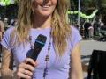 Una mujer con cabello largo y rizado, vestida con una camiseta púrpura y falda blanca, sostiene un micrófono. Fondo con decoración navideña y personas en el fondo.