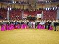 Una imagen de una ceremonia formal en un estadio con asientos rojos y amarillos. En el centro, una fila de personas vestidas con trajes formales y falda larga rosa. En el fondo, una pantalla de televisión, carteles con texto y un escenario con luces.