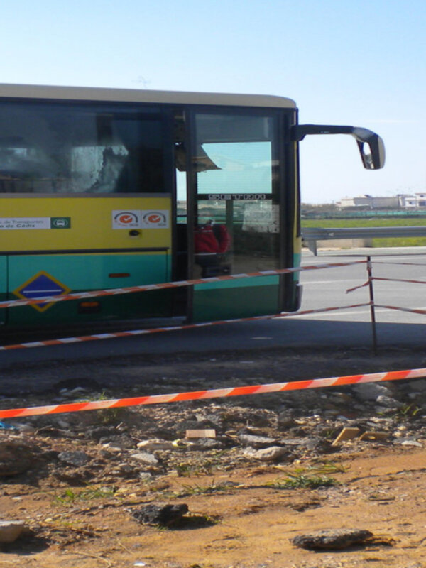 Un autobús amarillo y verde está estacionado en un área con una cerca roja y blanca. La imagen muestra el frente del vehículo, incluyendo la puerta y el espejo lateral. La cerca indica que hay algún tipo de restricción o precaución en el área, posiblemente debido a un trabajo de mantenimiento o una emergencia.