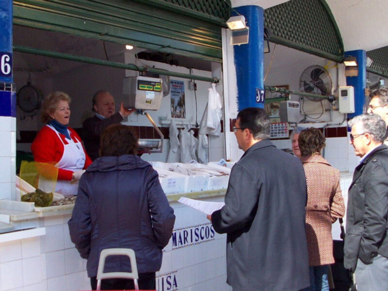 Un puesto de venta de mariscos con una mujer vendiendo pescado y un hombre comprando. El puesto tiene una señal que dice 'Mariscos' y está decorado con azulejos azules y blancos. Los clientes están esperando para comprar sus pescados frescos.