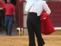Un torero en traje blanco y negro, con una capa roja, realiza un gesto de salto en la arena de toros. La imagen captura el momento de una corrida de toros, con un fondo borroso que muestra a otros espectadores y elementos típicos de una plaza de toros.